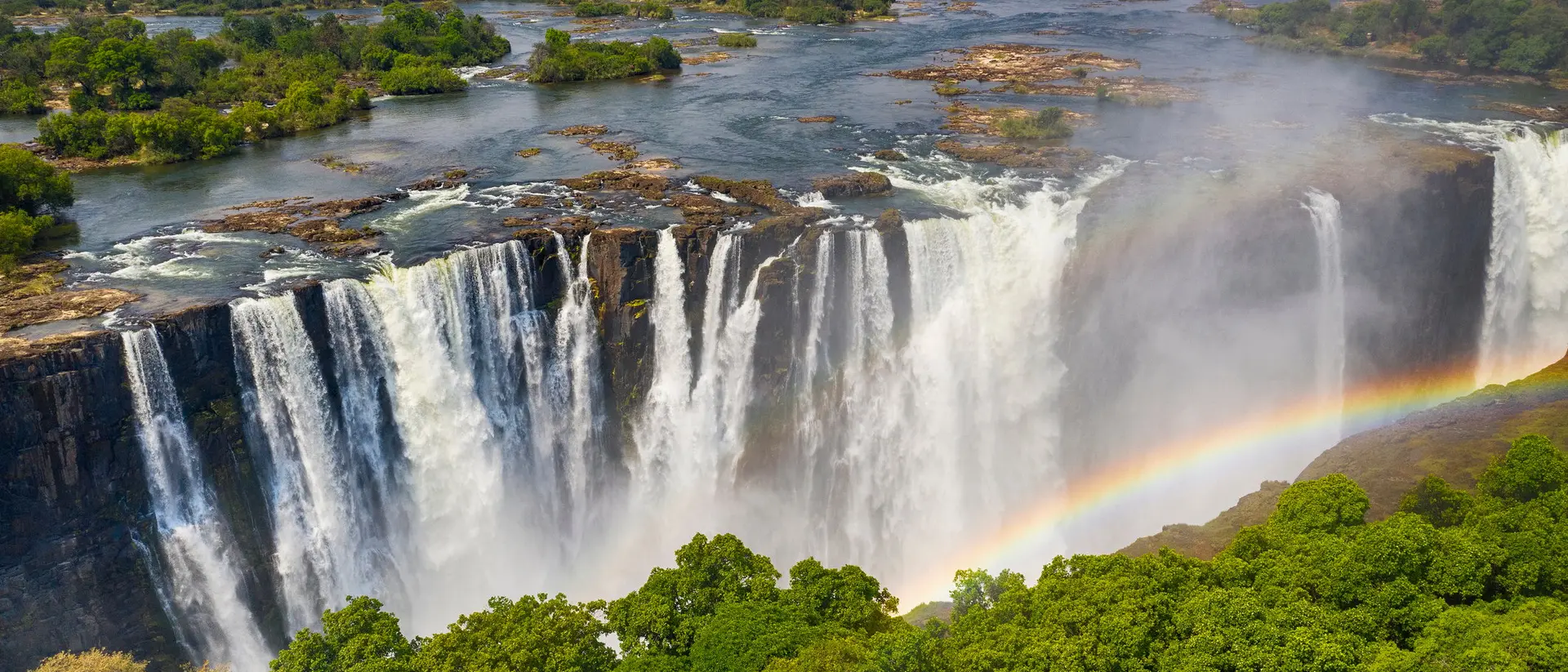 Aerial view of a majestic waterfall with a rainbow, surrounded by lush greenery and a flowing river.
