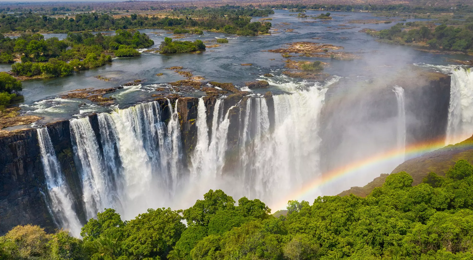Aerial view of a majestic waterfall with a rainbow, surrounded by lush greenery and a flowing river.