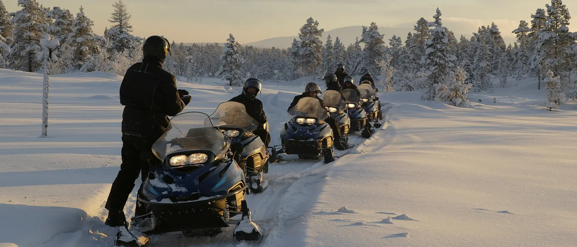 A group of snowmobilers riding through a snowy landscape with trees in the background during sunset.