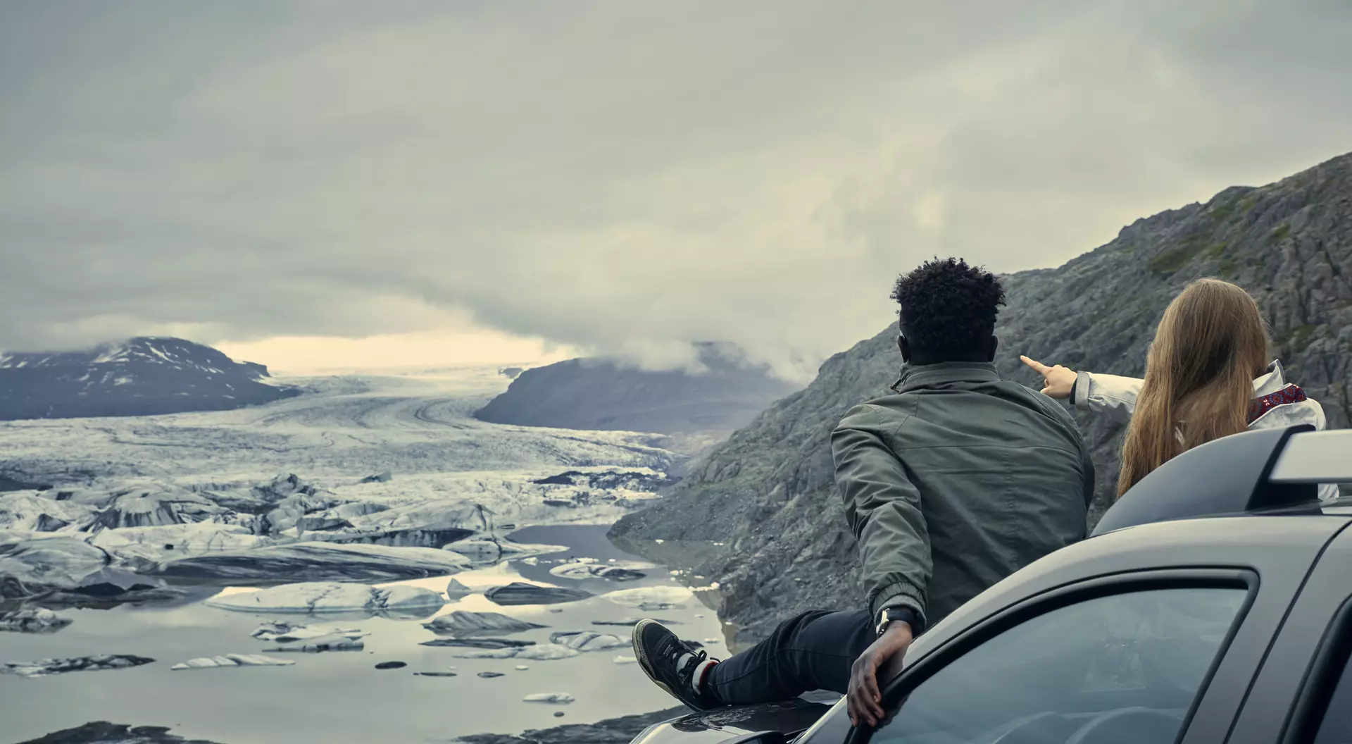 A couple sits on a car, gazing at a glacier landscape under a cloudy sky, pointing and enjoying the view.