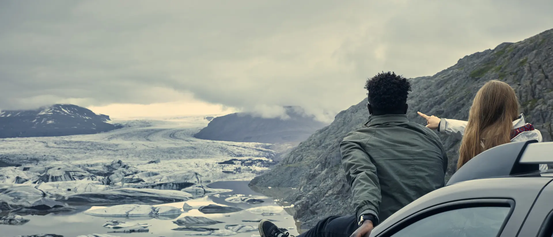 A couple sits on a car, gazing at a glacier landscape under a cloudy sky, pointing and enjoying the view.