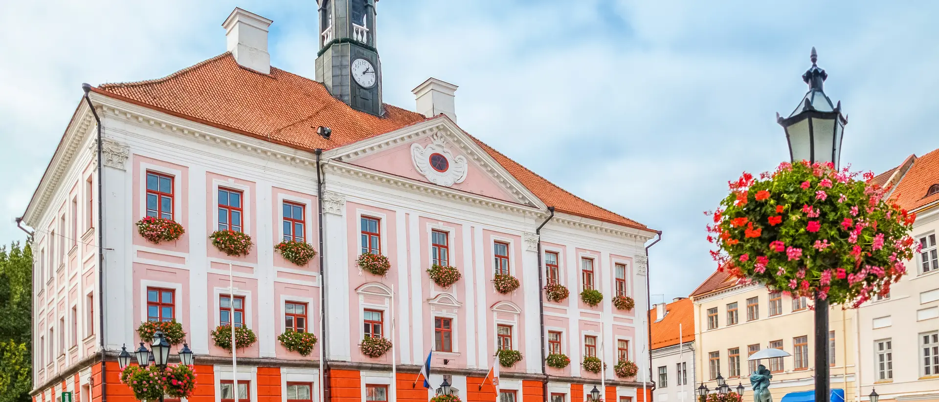 Pink and white town hall with a tower, surrounded by blooming planters and a cobblestone square on a cloudy day.