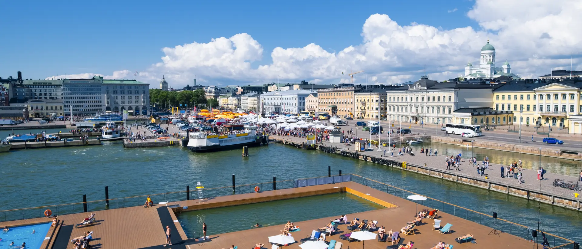View of a sunny harbor with people lounging on a deck, boats in the water, and buildings lining the shore.