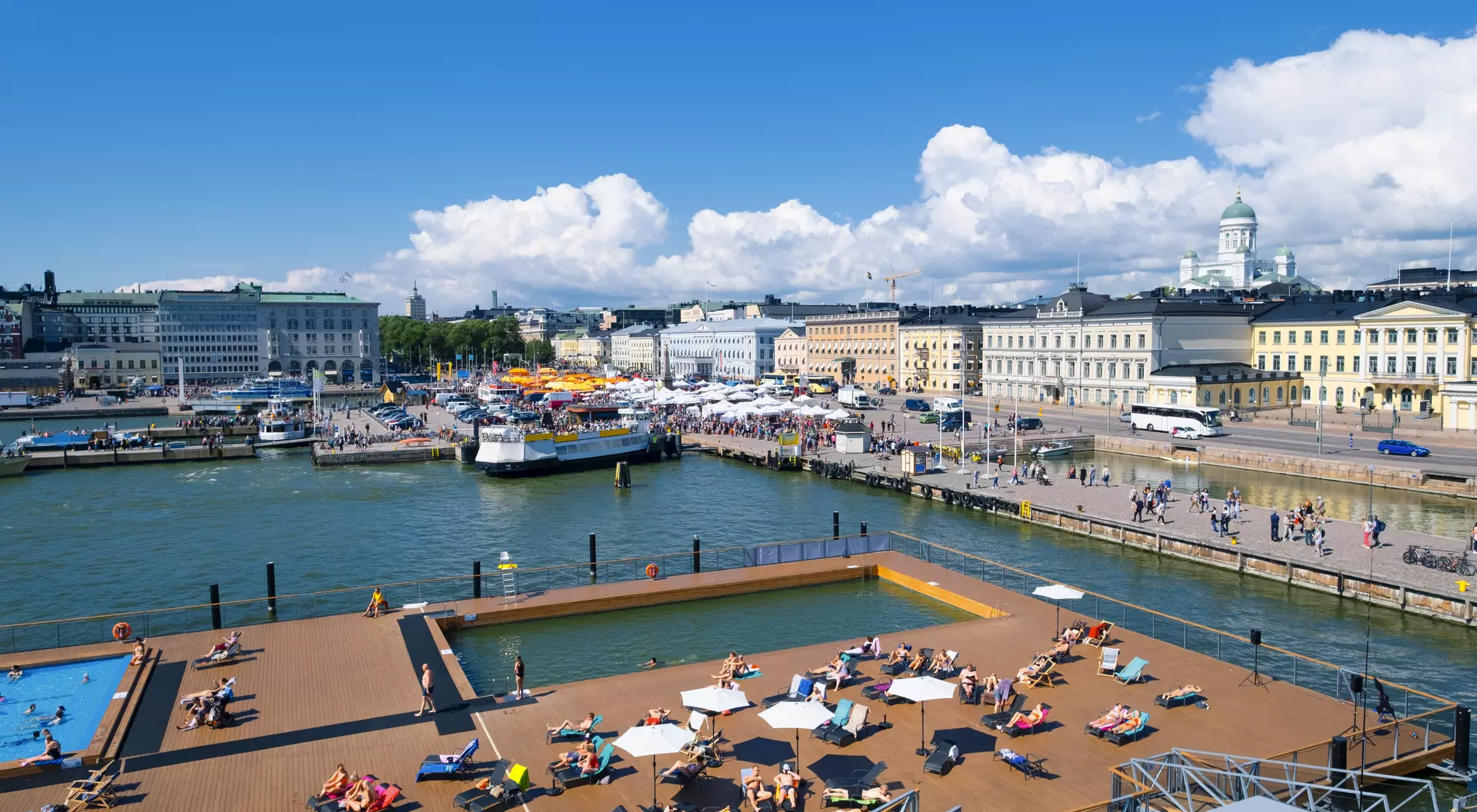 View of a sunny harbor with people lounging on a deck, boats in the water, and buildings lining the shore.