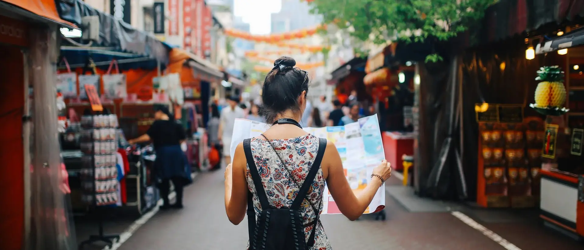 A woman stands in a market holding a map, surrounded by colorful stalls and hanging lanterns.