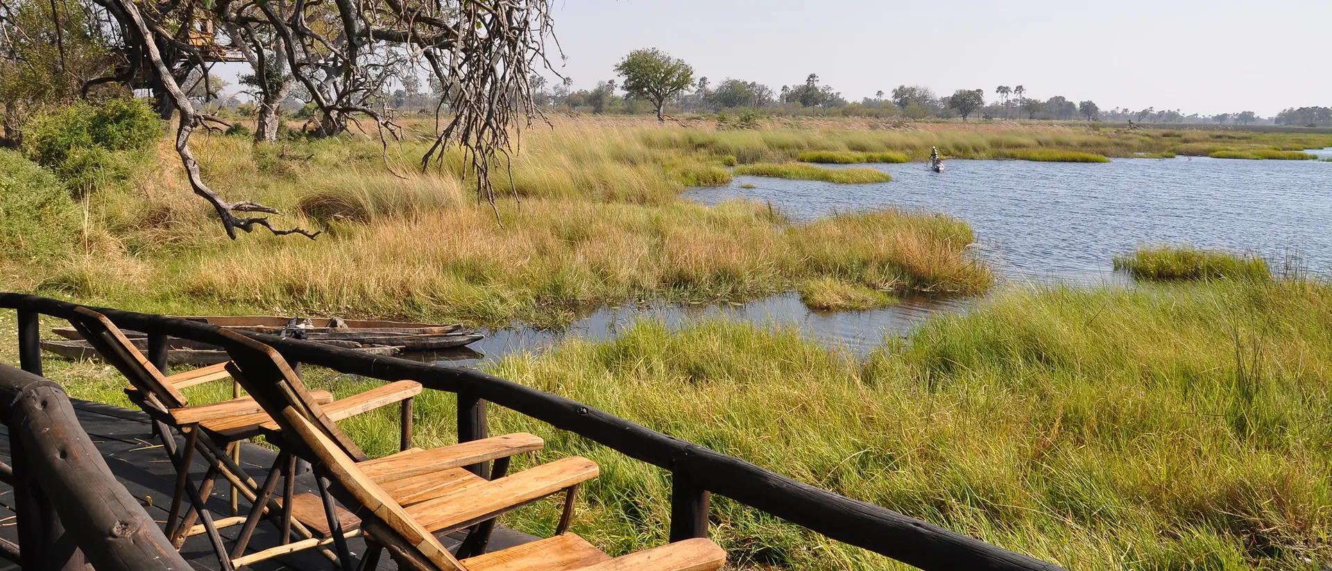 Wooden deck with empty chairs overlooks a serene wetland with lush grass and a tranquil waterway under a clear sky.