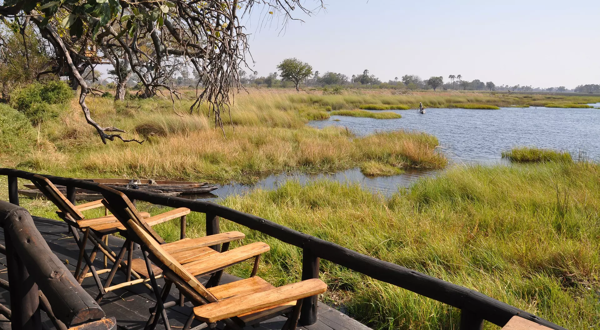 Wooden deck with empty chairs overlooks a serene wetland with lush grass and a tranquil waterway under a clear sky.