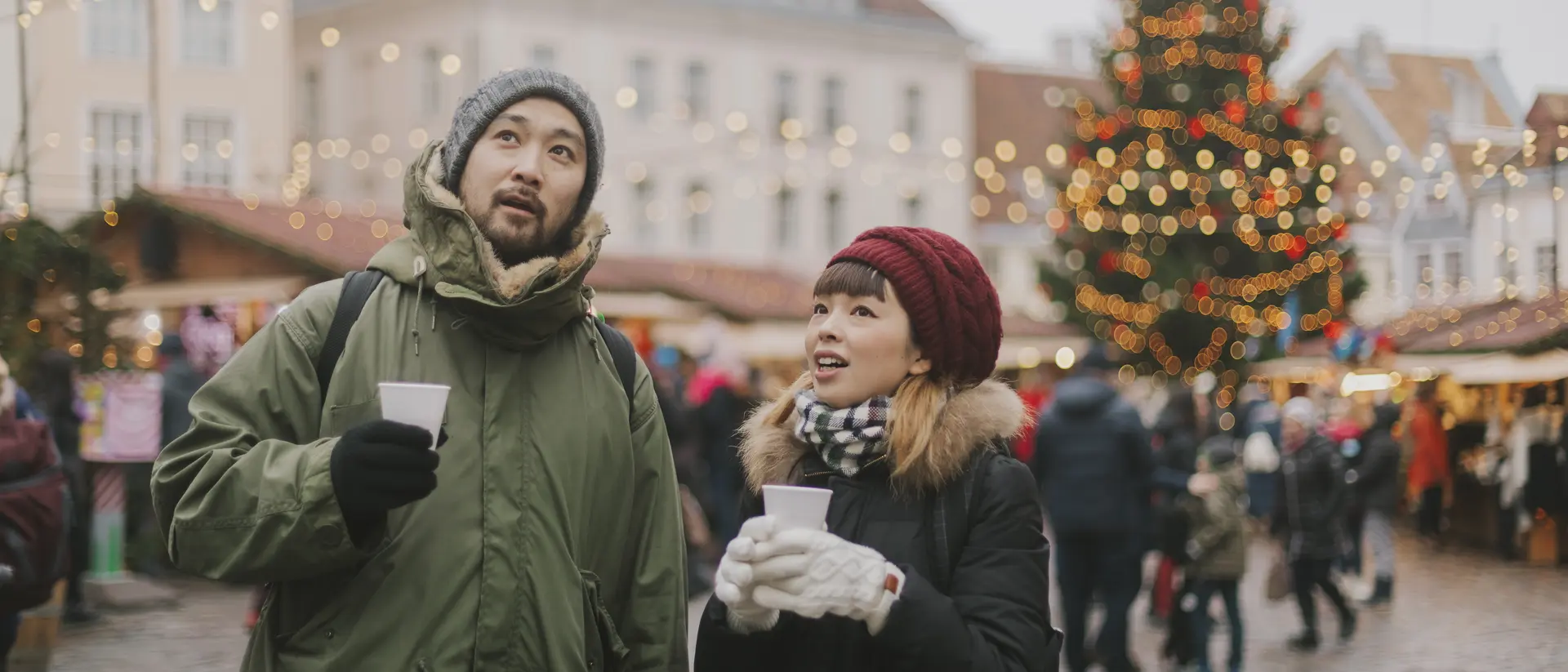 A man and woman look up, holding cups, in a festive market with holiday lights and a Christmas tree in the background.