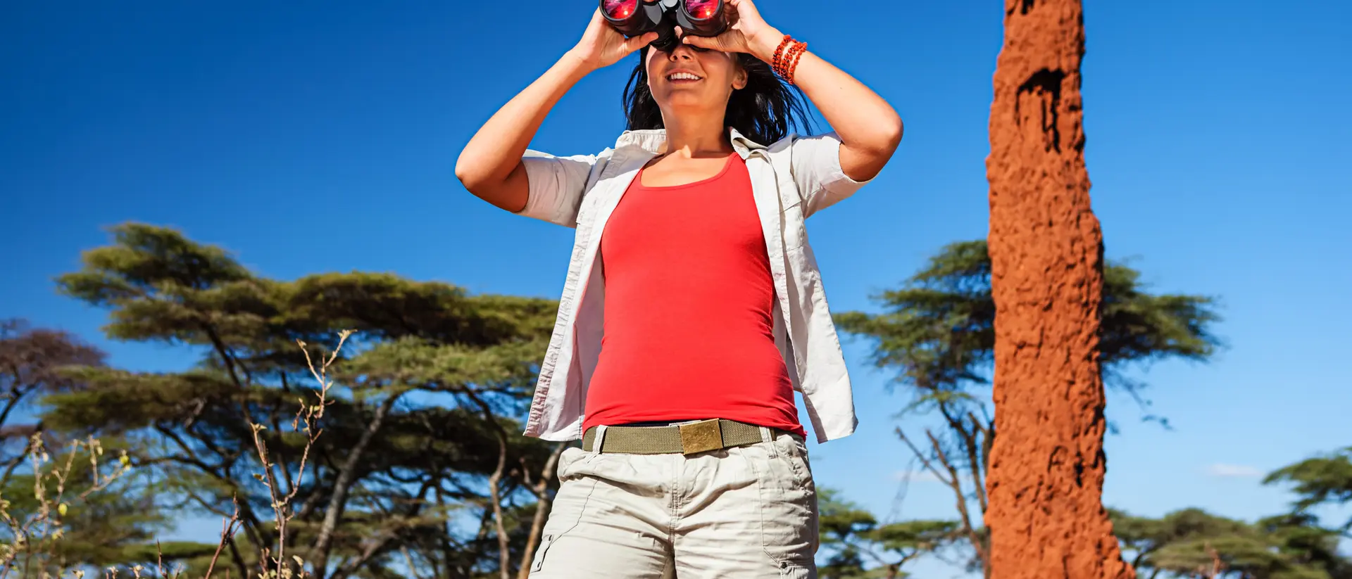 A woman in casual attire uses binoculars in a sunny landscape with distinctive red rock formations and sparse trees.