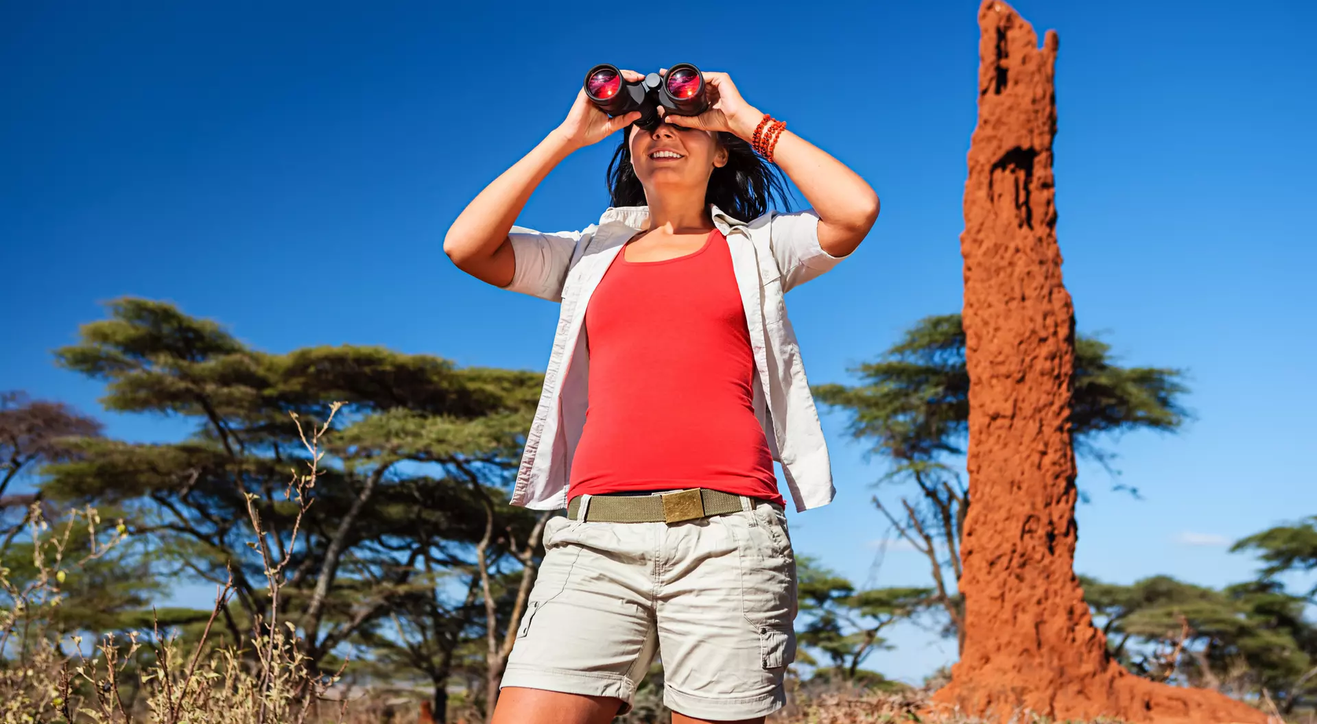 A woman in casual attire uses binoculars in a sunny landscape with distinctive red rock formations and sparse trees.