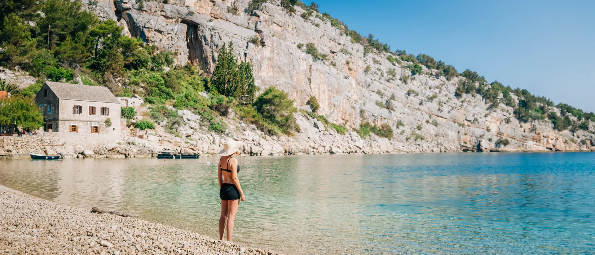 A person stands on a pebbled beach beside clear water, with rocky cliffs and a building in the background under a blue sky.