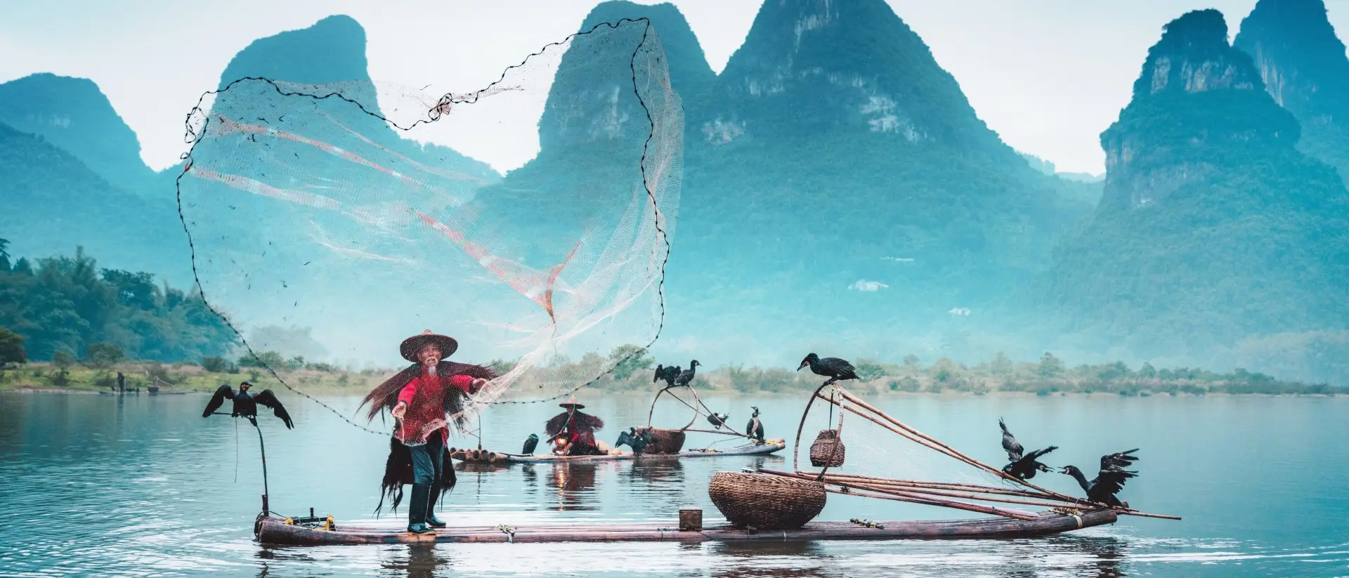 Fisherman on a bamboo raft casts a net, with cormorants perched nearby and misty mountains in the background.
