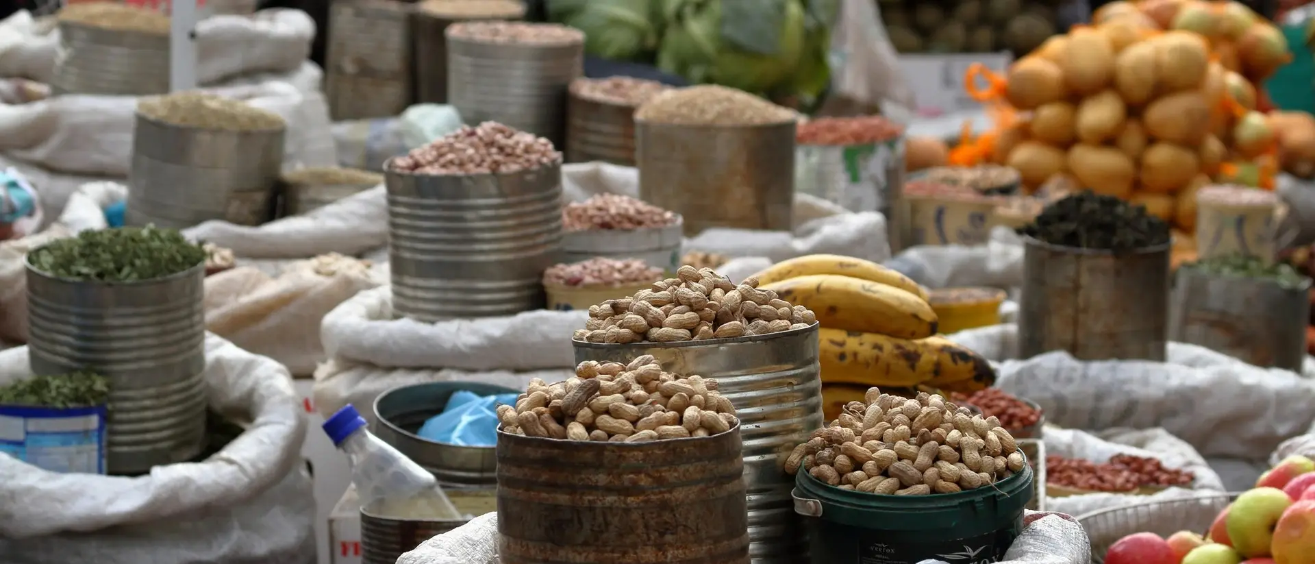 Various sacks of nuts and legumes are arranged in a bustling market, with fruits and vegetables in the background.