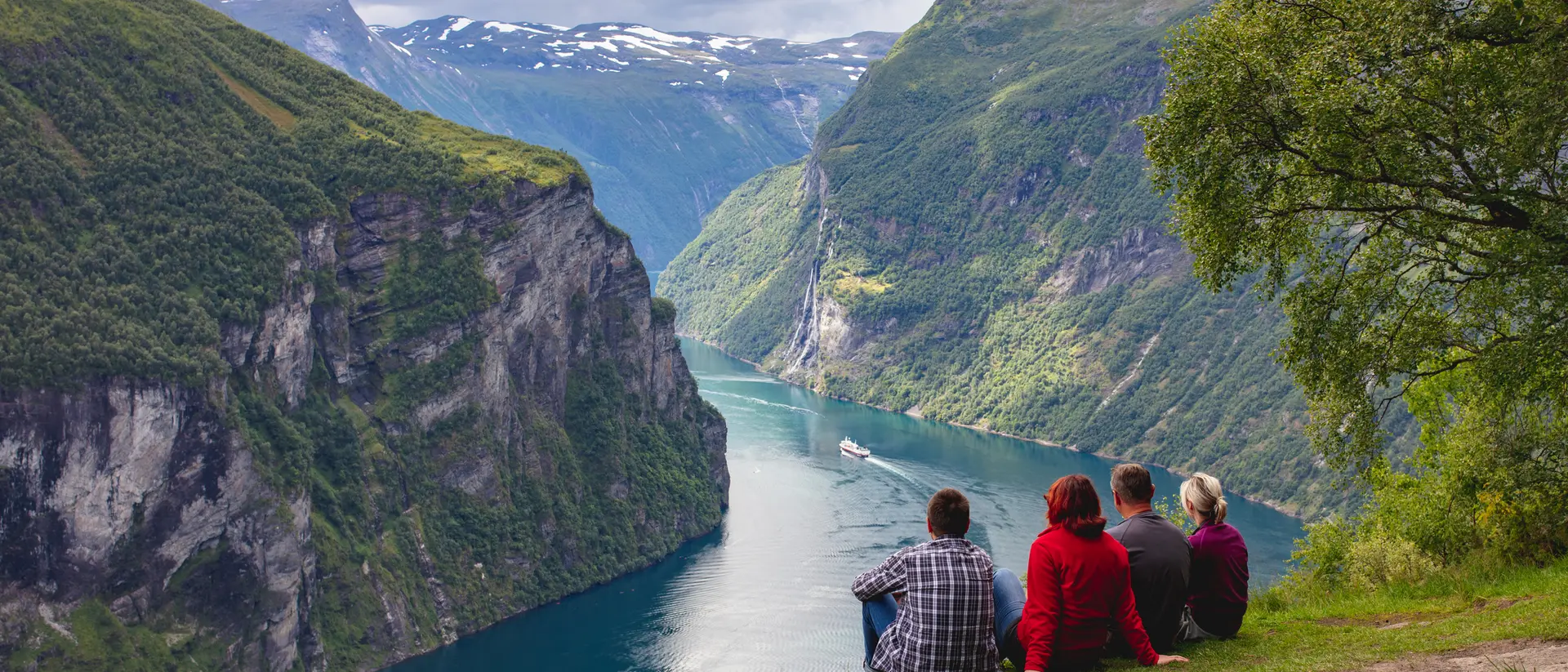 Four people sit on a ledge, overlooking a fjord surrounded by steep green cliffs and a calm blue waterway.