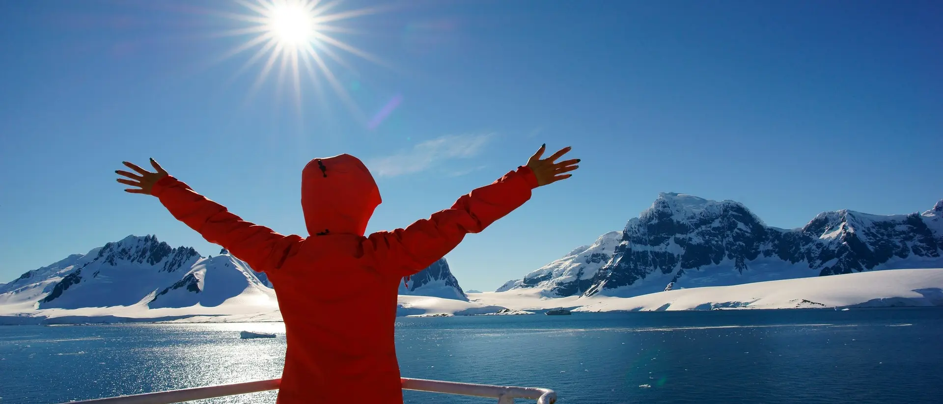 Person in a red jacket spreading arms wide, facing sunlit mountains and ocean under a clear blue sky.