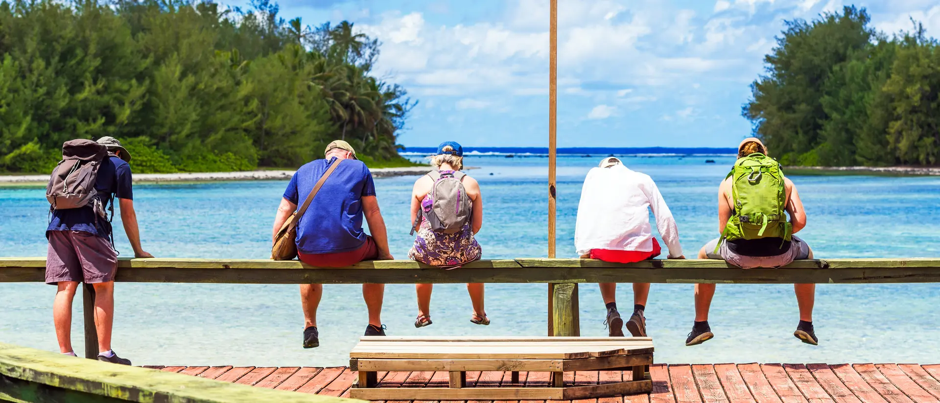 Five people sit on a wooden railing by the water, facing the ocean under a blue sky with scattered clouds.
