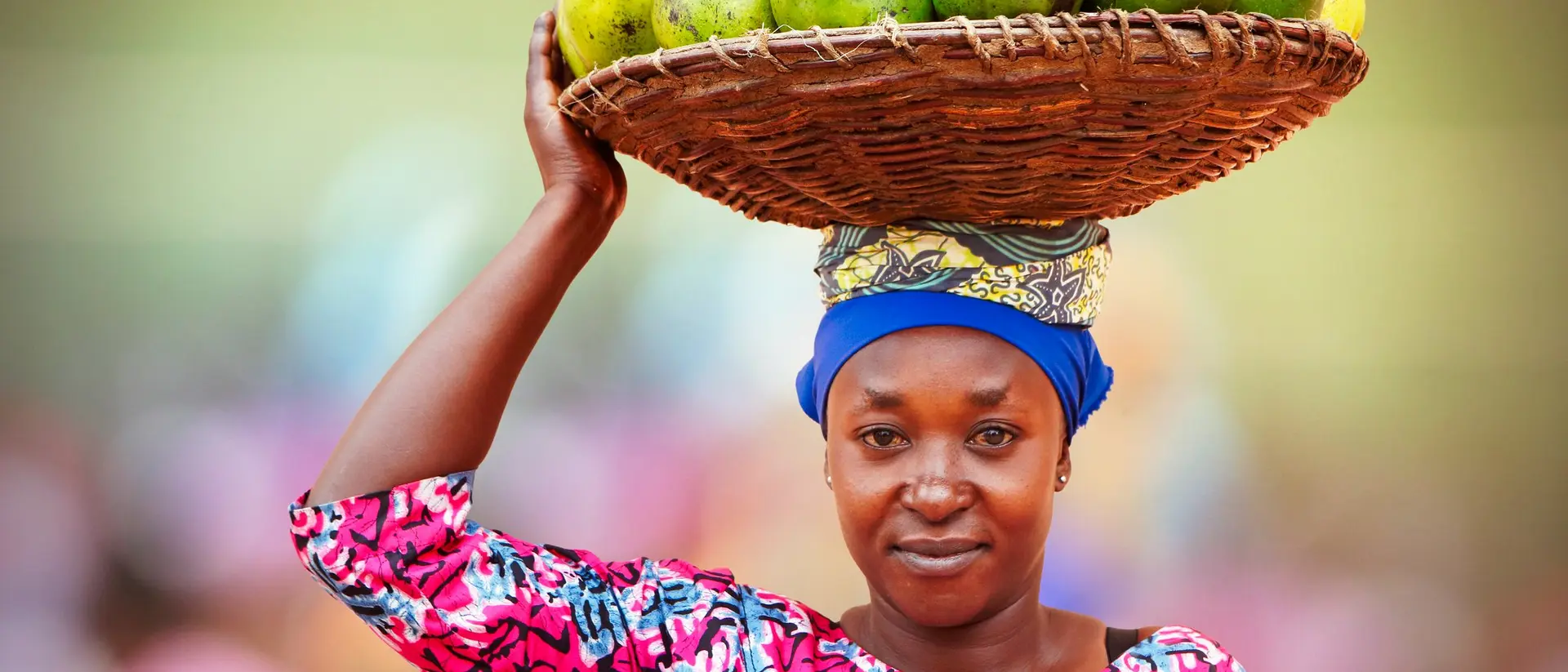 A woman in a colorful outfit balances a basket of fruits on her head while smiling at the camera.