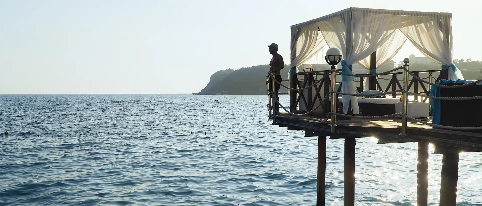 A person stands on a pier overlooking a calm ocean, with a tented seating area nearby and hills in the background.
