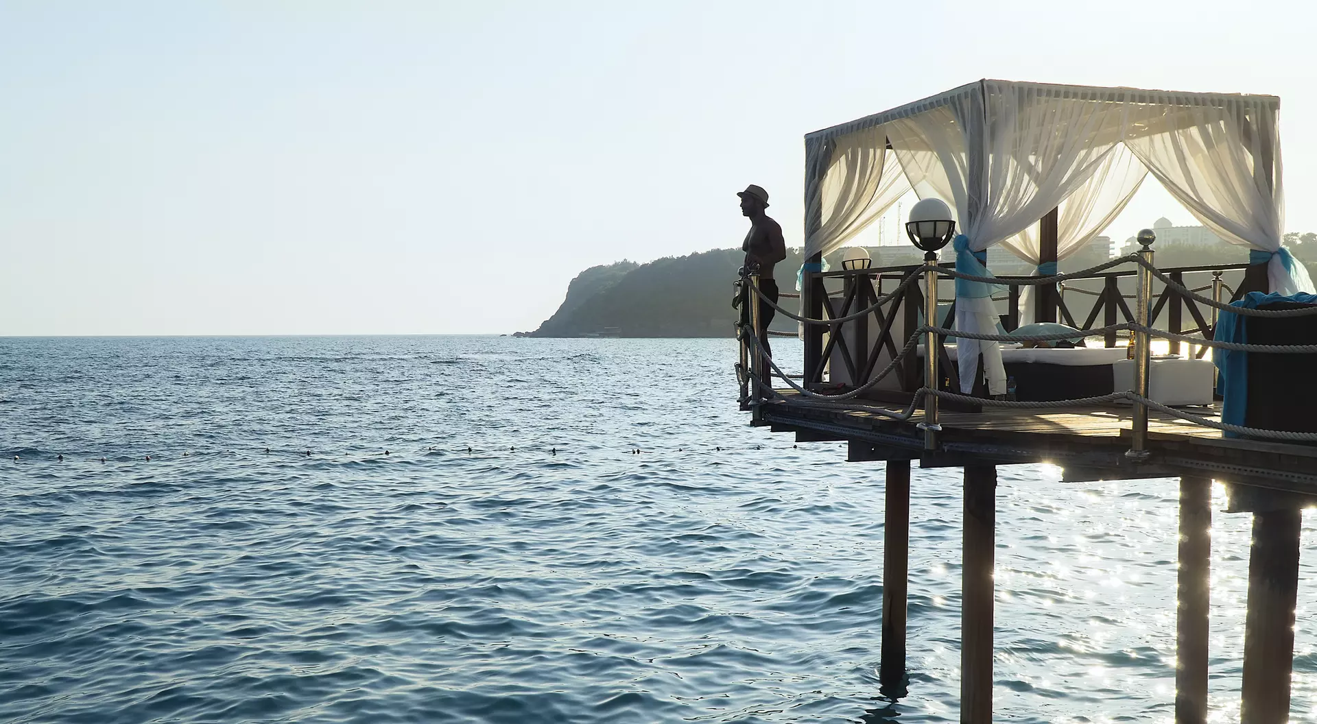 A person stands on a pier overlooking a calm ocean, with a tented seating area nearby and hills in the background.