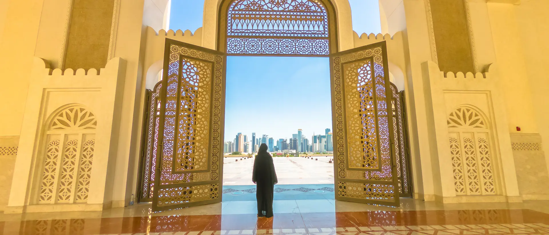 A person stands at an ornate doorway, looking out at a skyline through a bright, open arch.