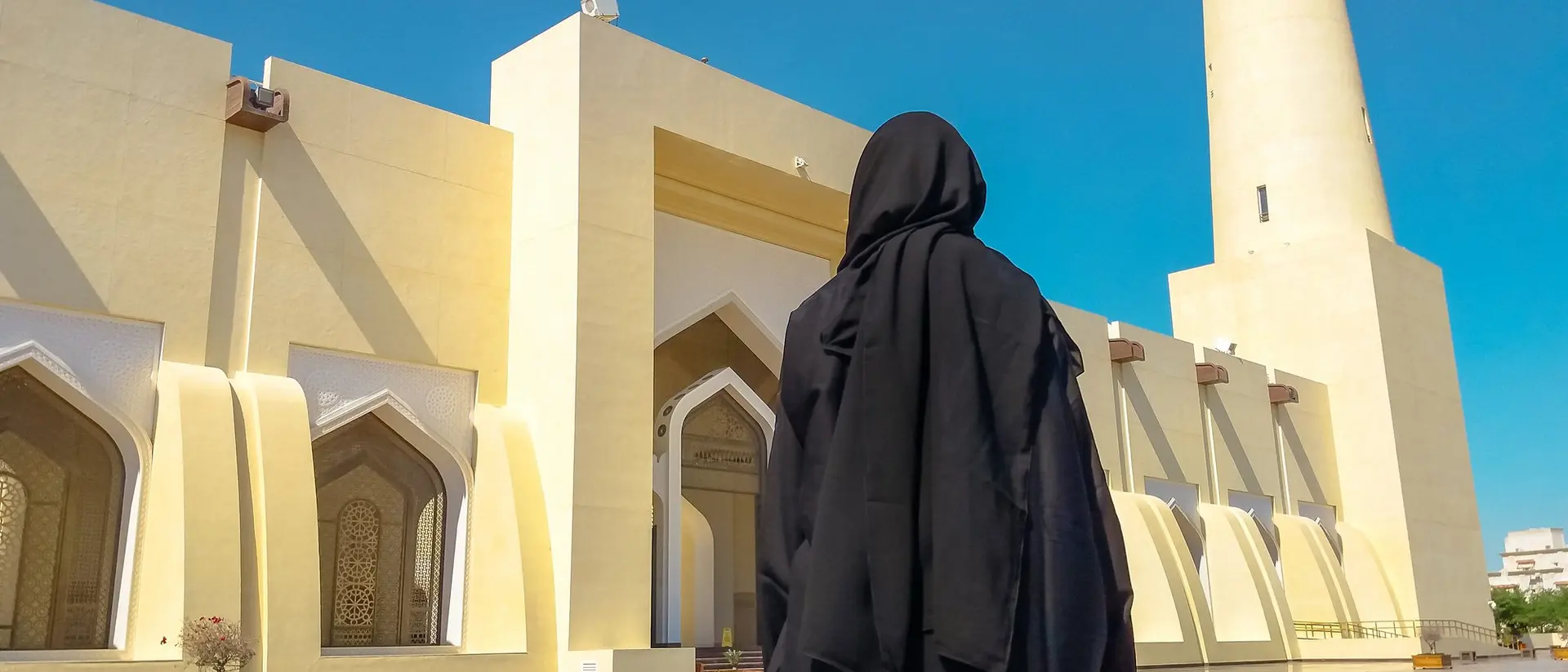 A person in a black abaya stands before a large mosque with a tall minaret under a blue sky.