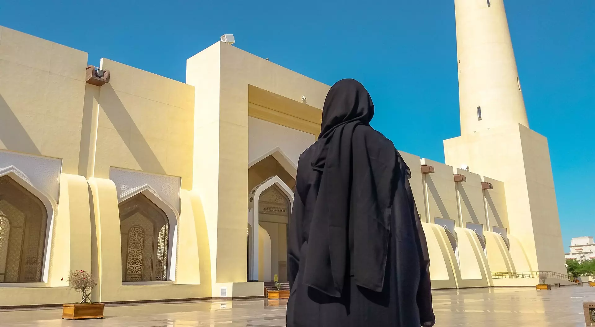 A person in a black abaya stands before a large mosque with a tall minaret under a blue sky.