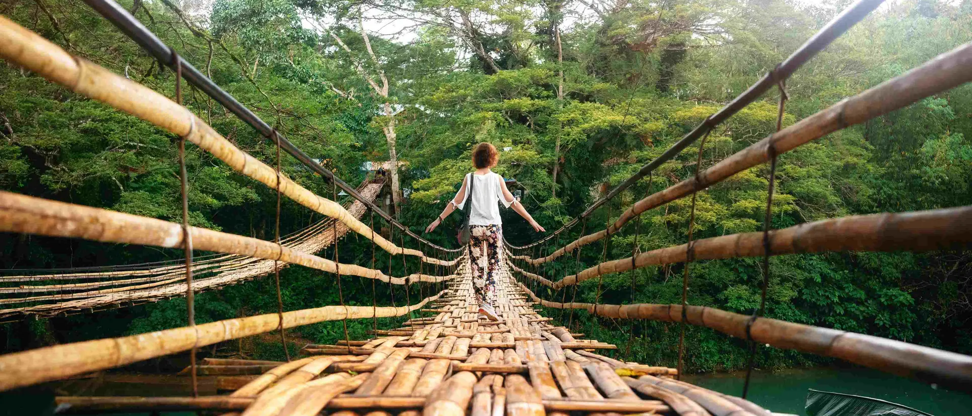 A child stands on a bamboo bridge surrounded by lush greenery, facing away from the camera.