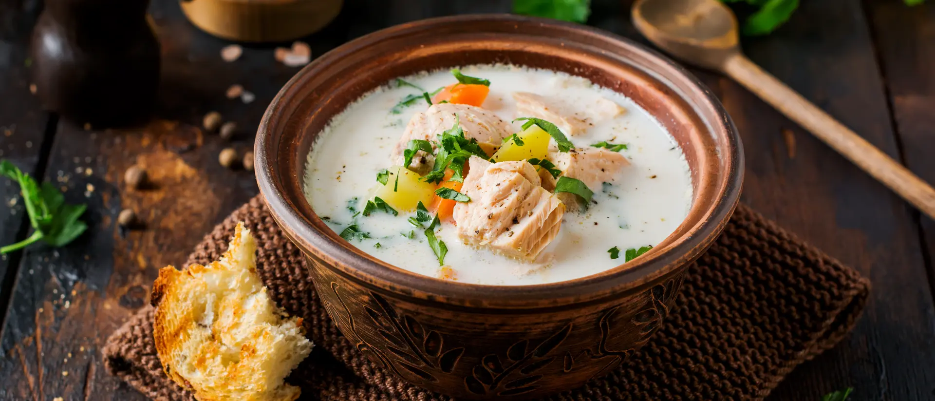 Bowl of creamy salmon soup with vegetables, garnished with herbs, beside a piece of bread on a woven mat.
