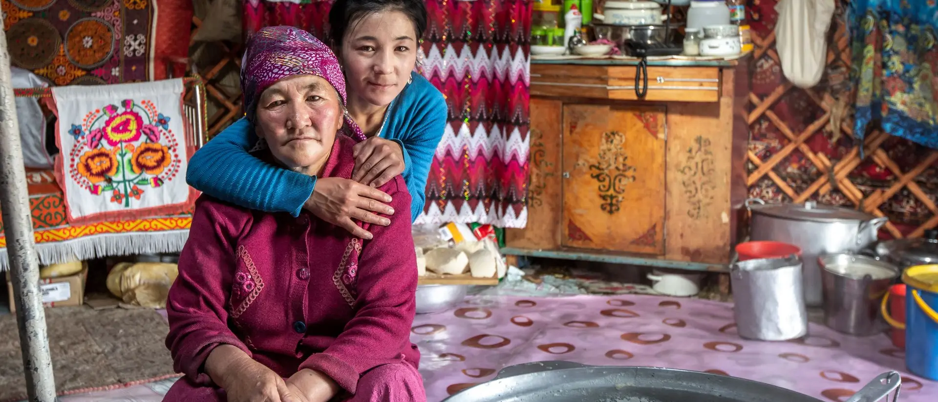 A young girl embraces an elderly woman in a cozy, traditional yurt adorned with colorful textiles and kitchen items.