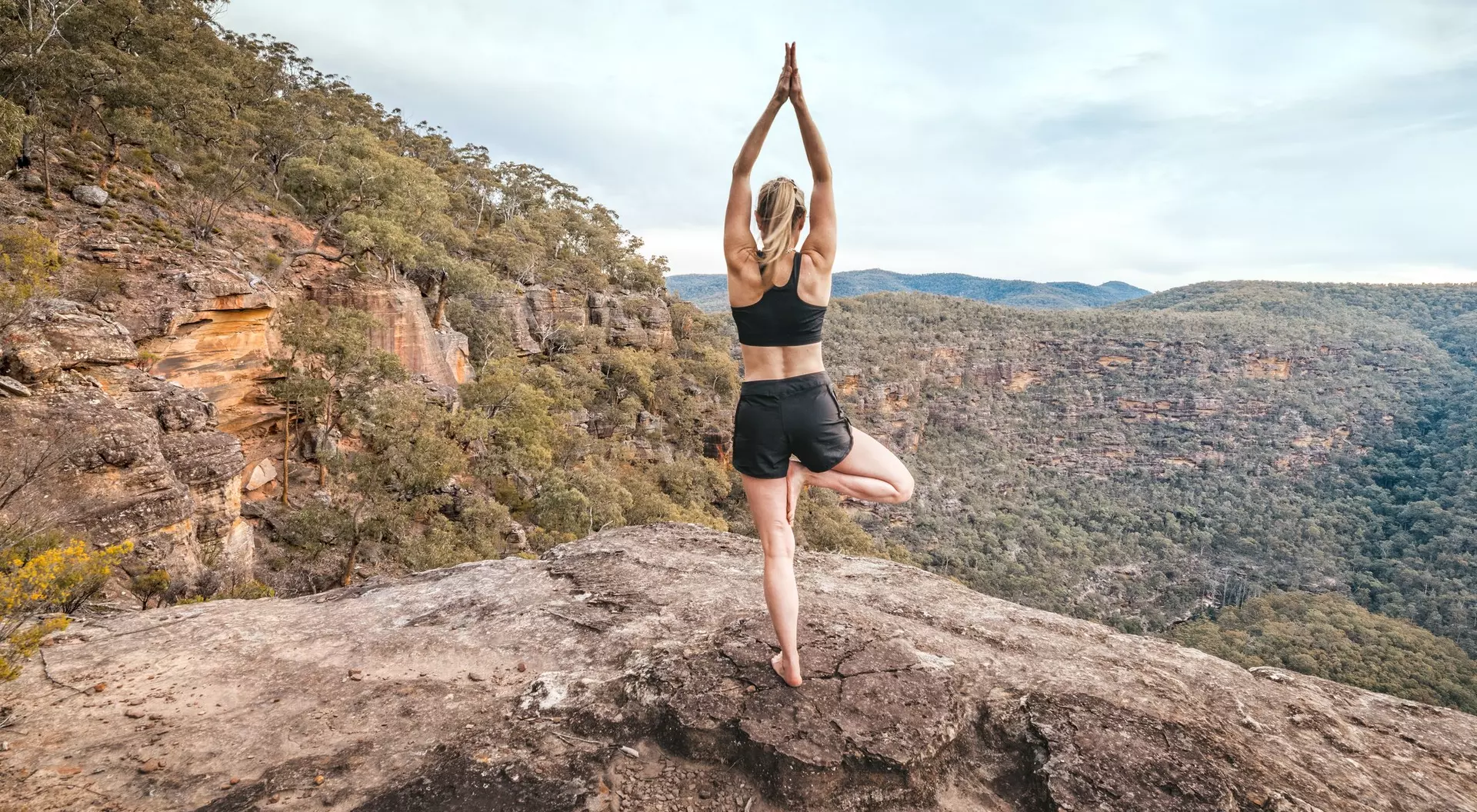 A person practicing yoga on one leg atop a rocky ledge, surrounded by trees and a scenic mountainous landscape.