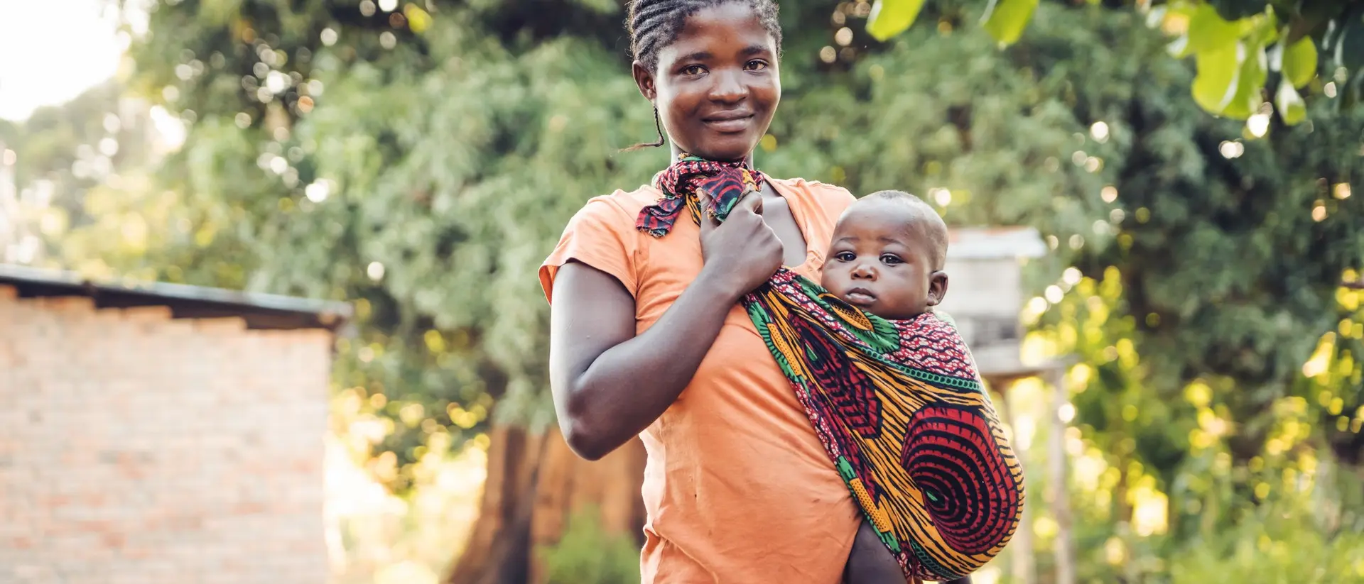 A woman wearing an orange top holds a baby wrapped in colorful fabric, standing outdoors near a home with trees in the background.
