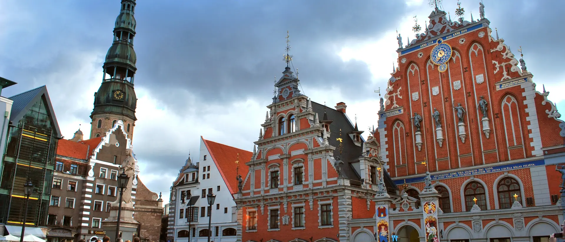 A bustling square featuring historic buildings, including a church tower and colorful merchant houses under a cloudy sky.
