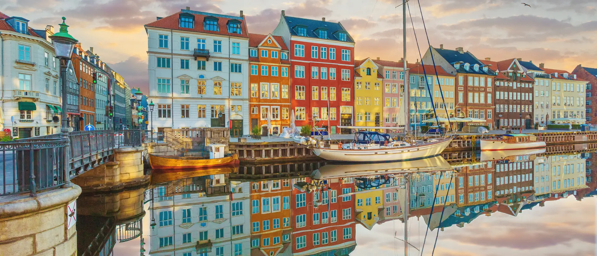 Colorful buildings line a canal at sunset, reflecting in the calm water, with a sailboat moored nearby.
