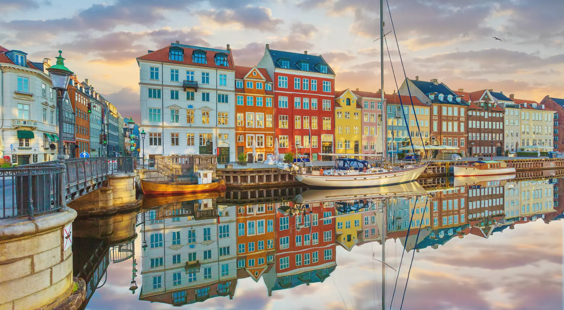 Colorful buildings line a canal at sunset, reflecting in the calm water, with a sailboat moored nearby.