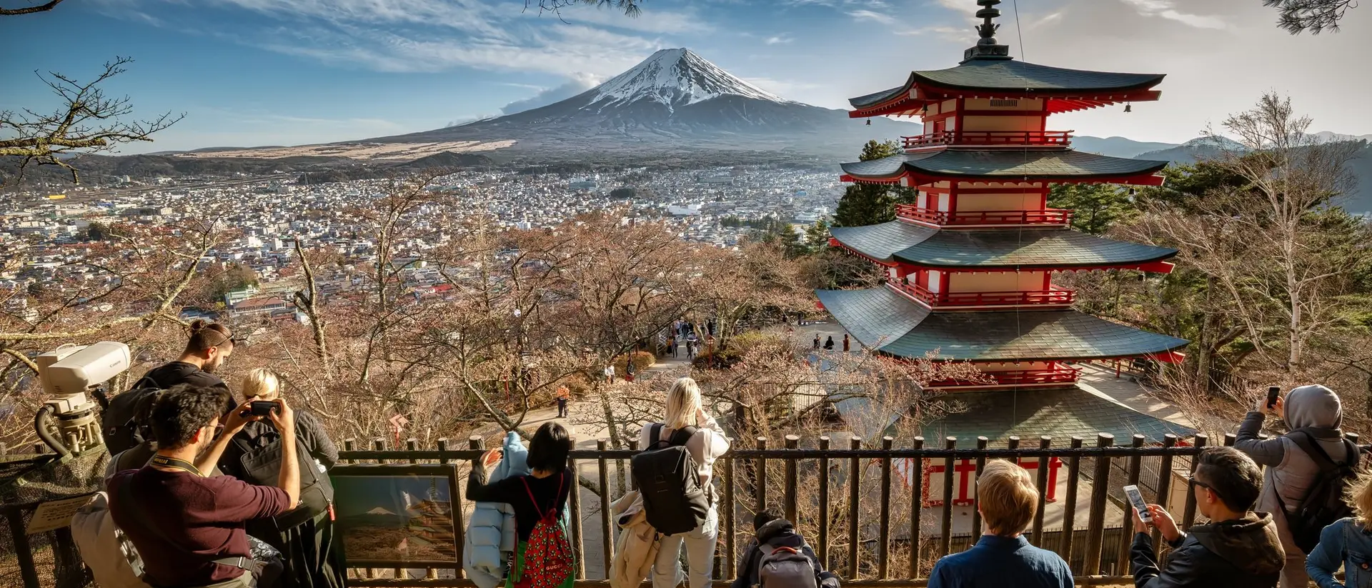 Viewers admire Mount Fuji and a red pagoda from a viewpoint, surrounded by trees on a clear day.