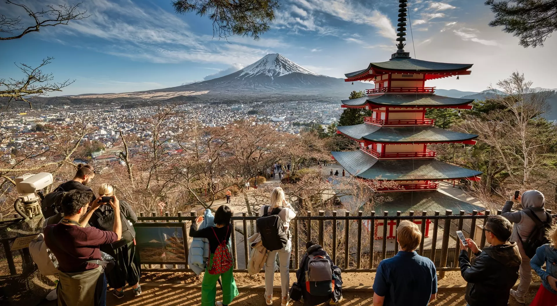 Viewers admire Mount Fuji and a red pagoda from a viewpoint, surrounded by trees on a clear day.
