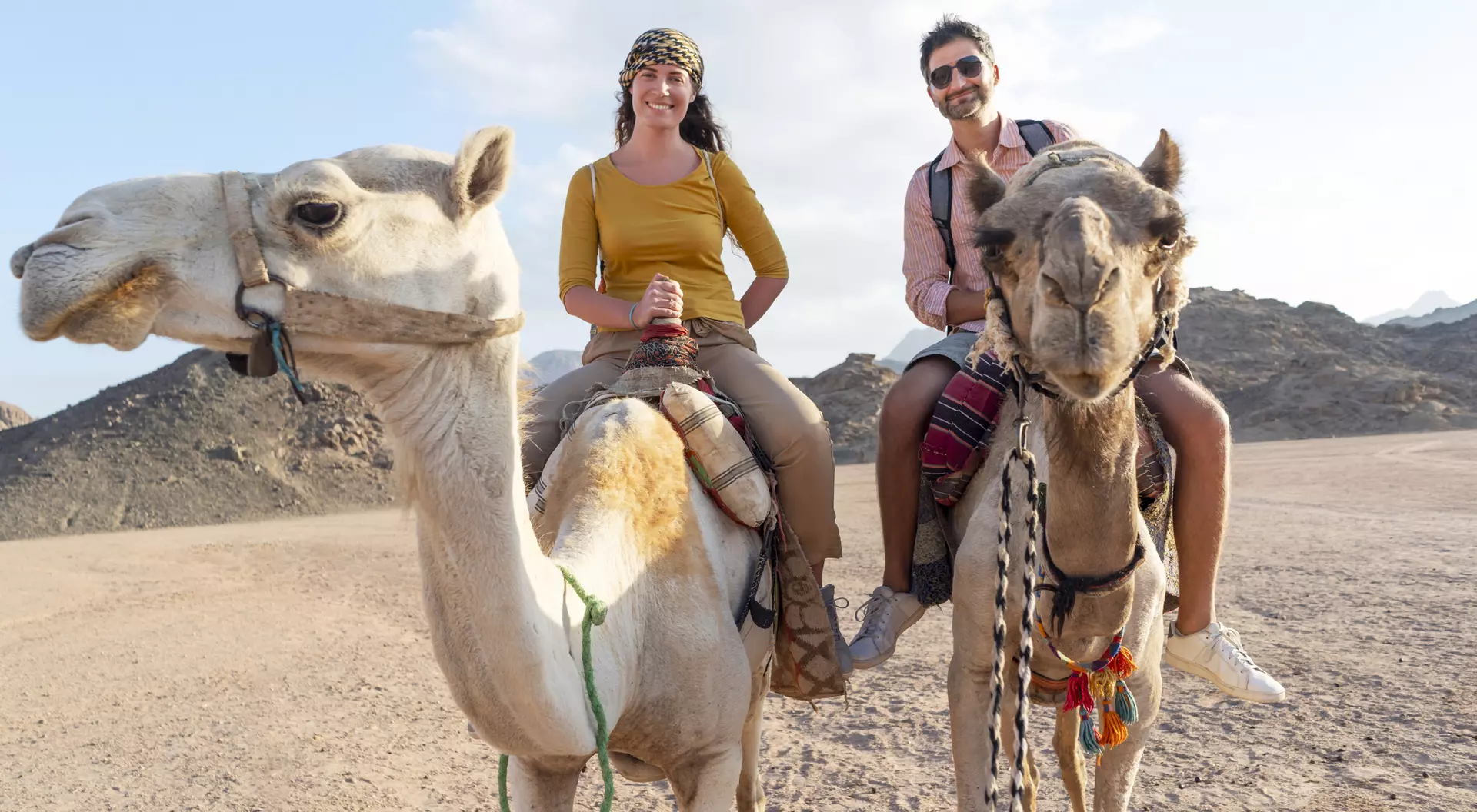 Two people ride camels in a desert landscape, smiling, with rocky mountains in the background.