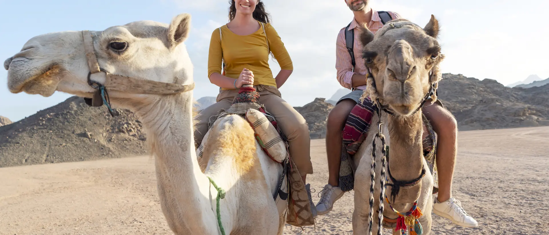 Two people ride camels in a desert landscape, smiling, with rocky mountains in the background.