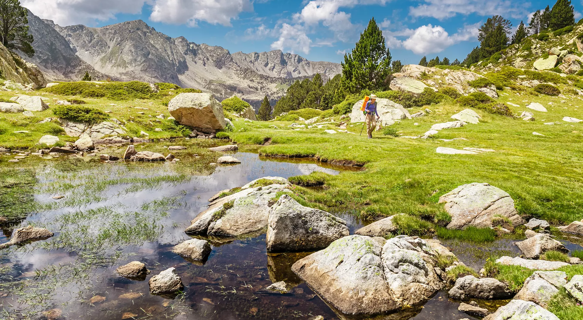 Hiker walking near a serene pond, surrounded by lush greenery and rocky terrain, with mountains in the background.