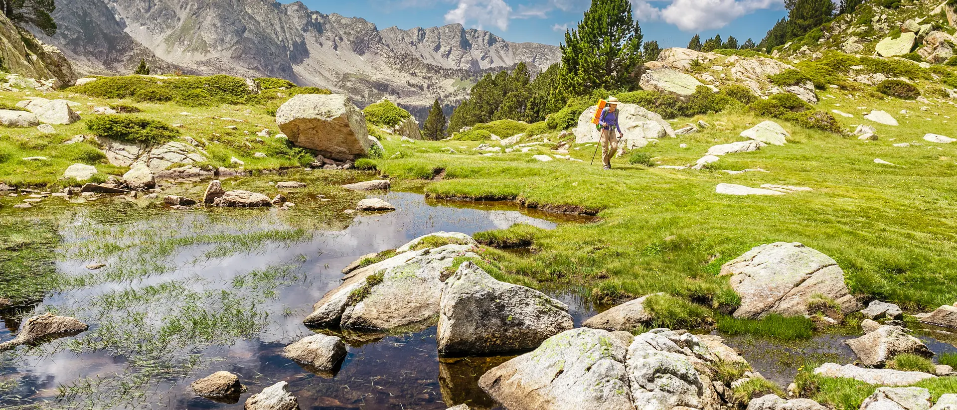 Hiker walking near a serene pond, surrounded by lush greenery and rocky terrain, with mountains in the background.