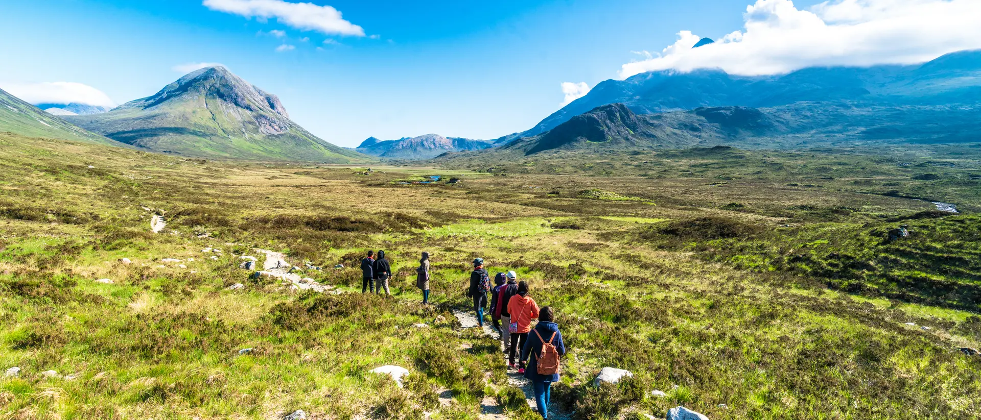 Group of hikers walking on a trail through a lush green landscape with mountains under a clear blue sky.