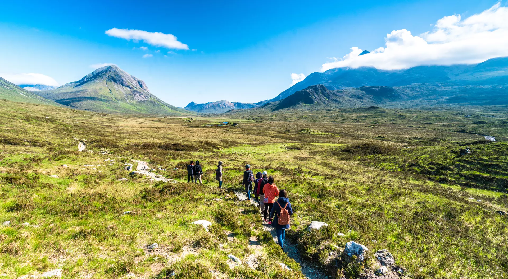 Group of hikers walking on a trail through a lush green landscape with mountains under a clear blue sky.