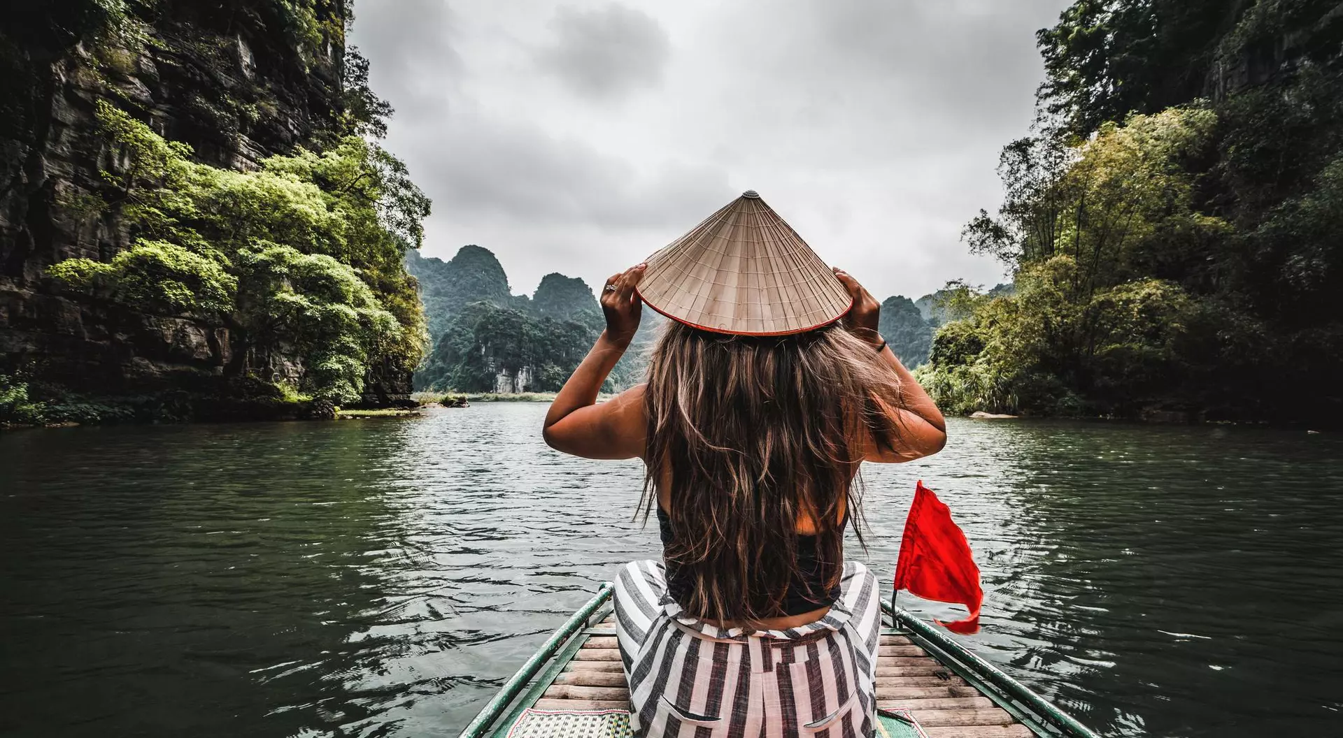 A person in a conical hat sits on a boat, gazing at scenic mountains and lush greenery on a calm lake.