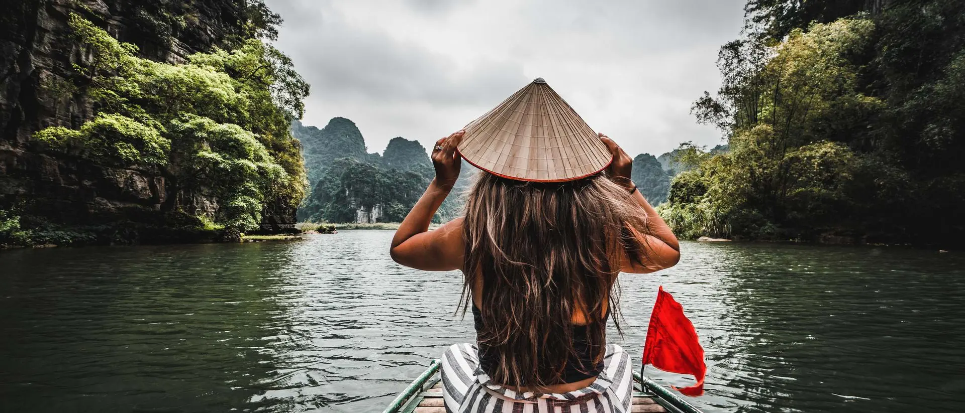 A person in a conical hat sits on a boat, gazing at scenic mountains and lush greenery on a calm lake.