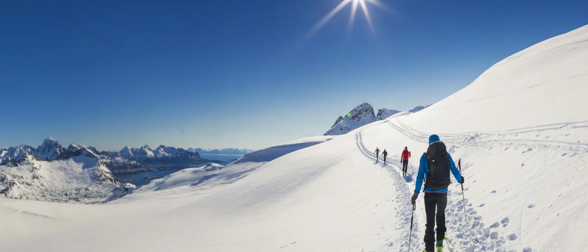 Four individuals ski across a snowy landscape under a bright sun and clear blue sky, with mountains in the background.