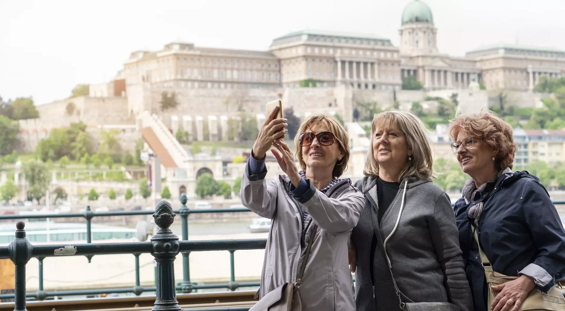 Three women take a selfie with a historic building and river in the background.
