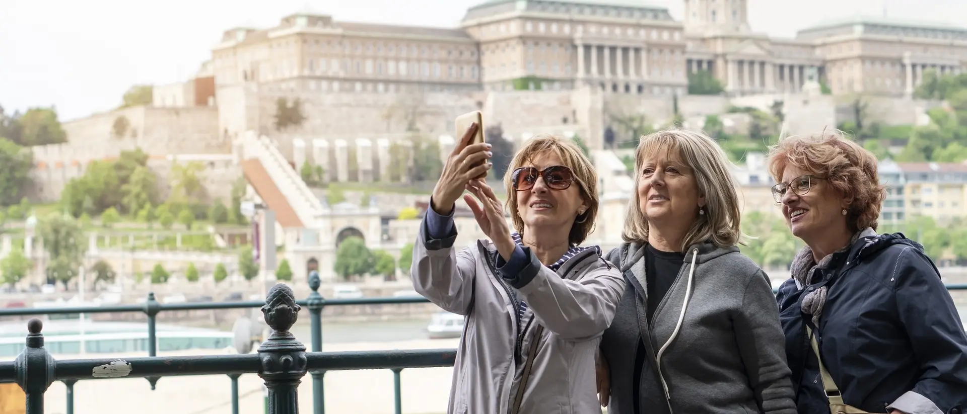Three women take a selfie with a historic building and river in the background.