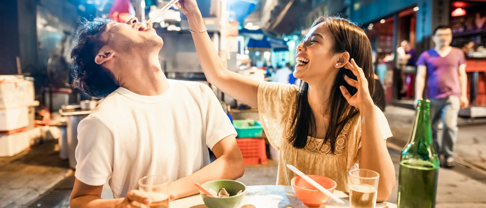 A woman playfully feeds a man while enjoying a meal at a lively outdoor food market.
