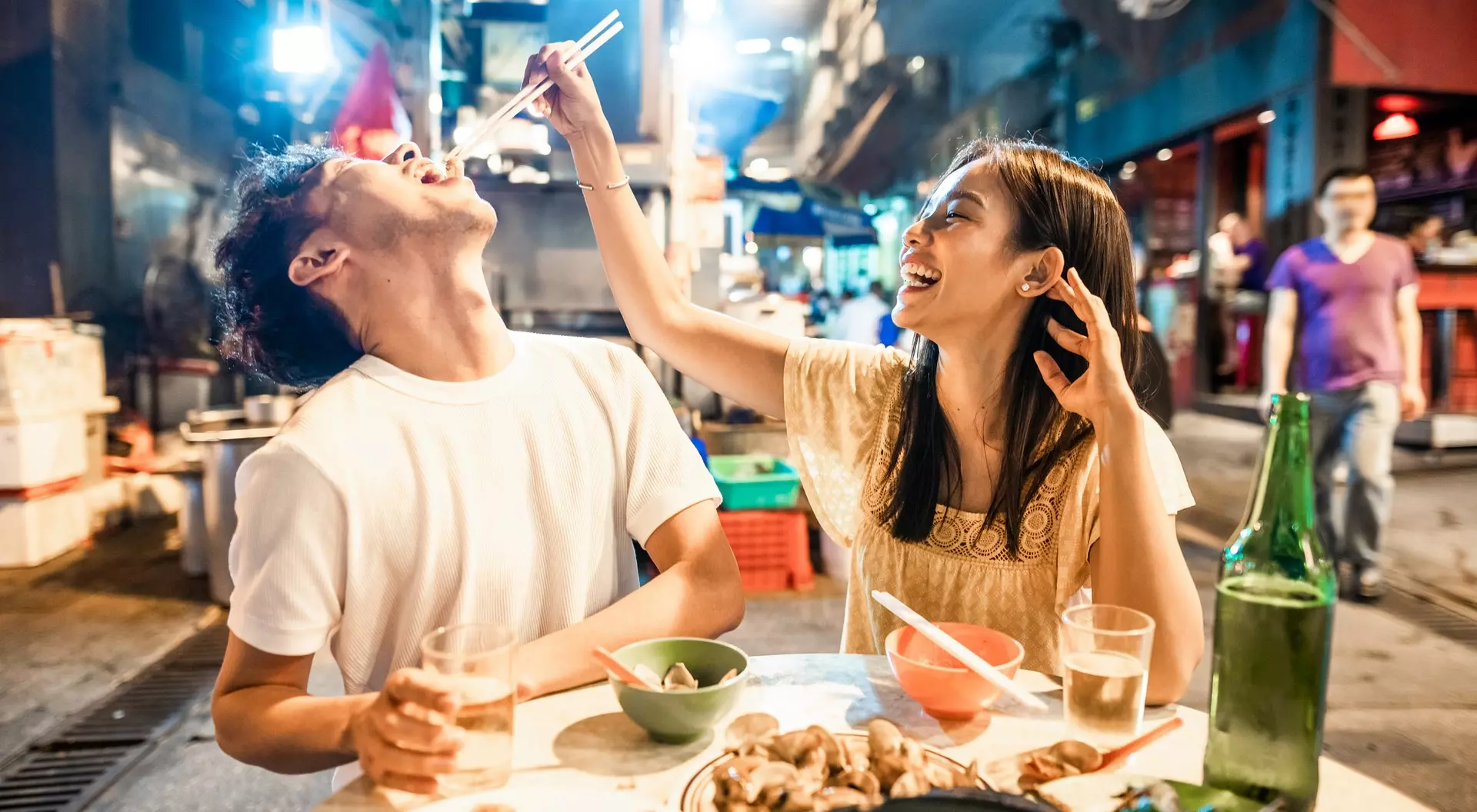 A woman playfully feeds a man while enjoying a meal at a lively outdoor food market.