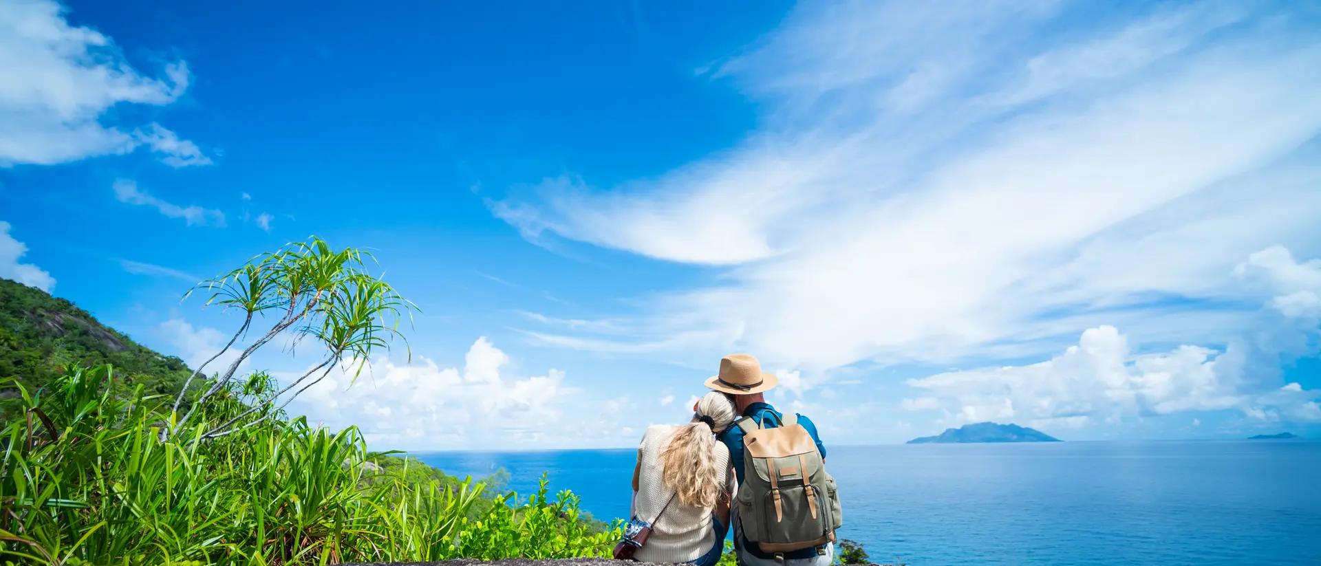 A couple sits on a rock, gazing at the ocean and blue sky, surrounded by greenery.